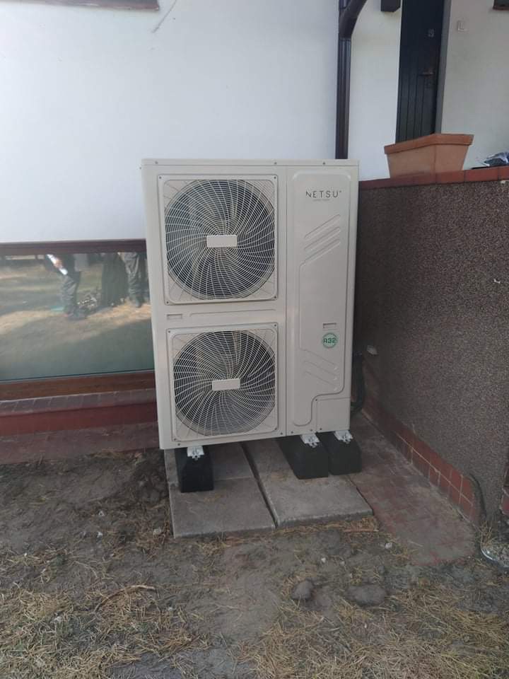 Biała, dwuczęściowa pompa ciepła Netsu, zamontowana na betonowych płytach przed budynkiem z widocznym oknem i rynną, na tle ziemi z resztkami trawy.