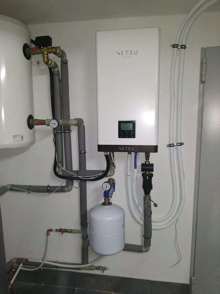 Biały, wiszący magazyn energii Netsu z podłączonymi rurami i zbiornikiem wyrównawczym, zamontowany na jasnej ścianie w pomieszczeniu technicznym.