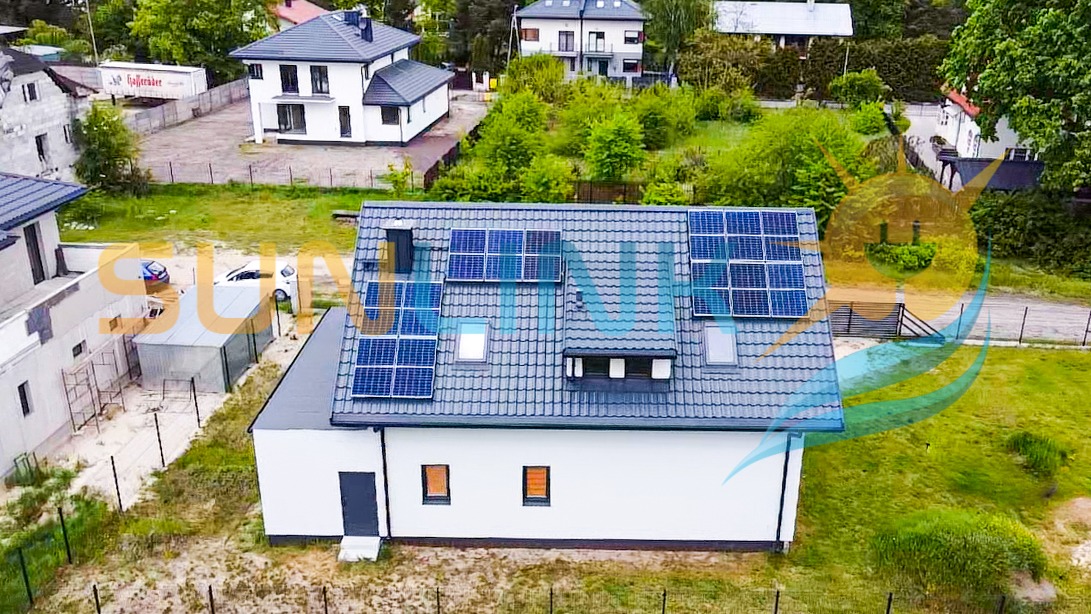 Biały dom jednorodzinny z szarą dachówką, na której zamontowano panele słoneczne. Widok z góry, w tle zieleń i inne budynki.