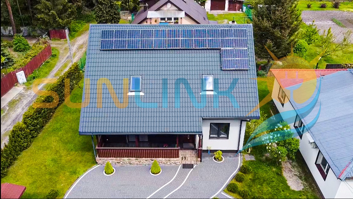 Dom jednorodzinny z szarym dachem i panelami słonecznymi, widok z góry. Uwagę przykuwają starannie przystrzyżone krzewy i kostka brukowa na podjeździe.