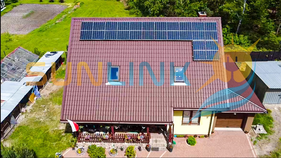 Dom jednorodzinny z brązowym dachem i panelami słonecznymi na połaci. Widok z lotu ptaka, otoczenie zielone, z boku garaż. Polska flaga na balkonie.