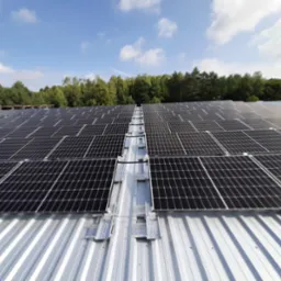 Rozległa instalacja fotowoltaiczna na metalowym dachu budynku przemysłowego, widok z góry, zalesione tło pod błękitnym niebem z chmurami.