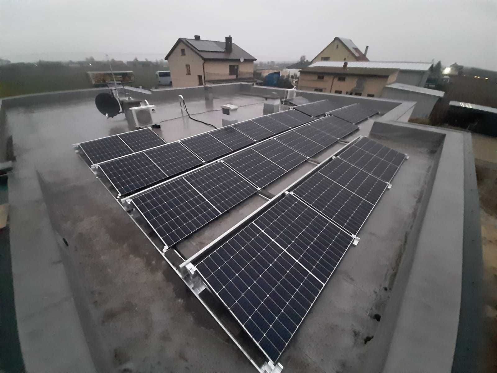 Rząd paneli słonecznych na płaskim dachu budynku mieszkalnego w pochmurny dzień, widoczny również klimatyzator zewnętrzny i antena satelitarna.