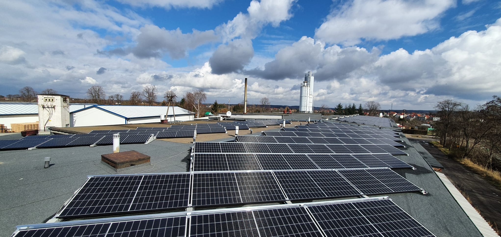 Rozległa instalacja fotowoltaiczna na dachu budynku przemysłowego, widok z góry, błękitne niebo z chmurami w tle.
