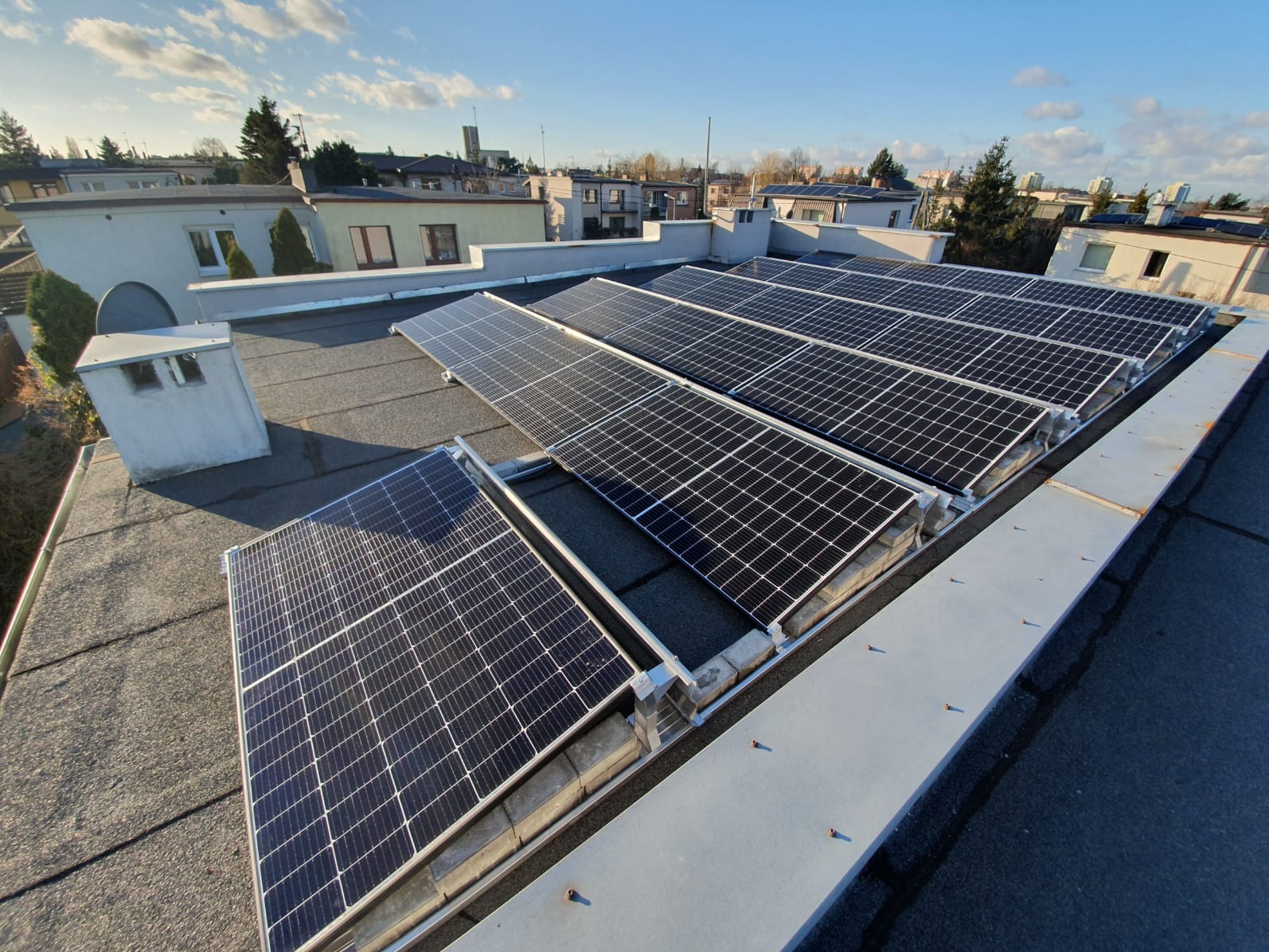 Panele słoneczne zamontowane na płaskim dachu budynku mieszkalnego, widok z góry na tle zabudowy miejskiej i błękitnego nieba z chmurami.