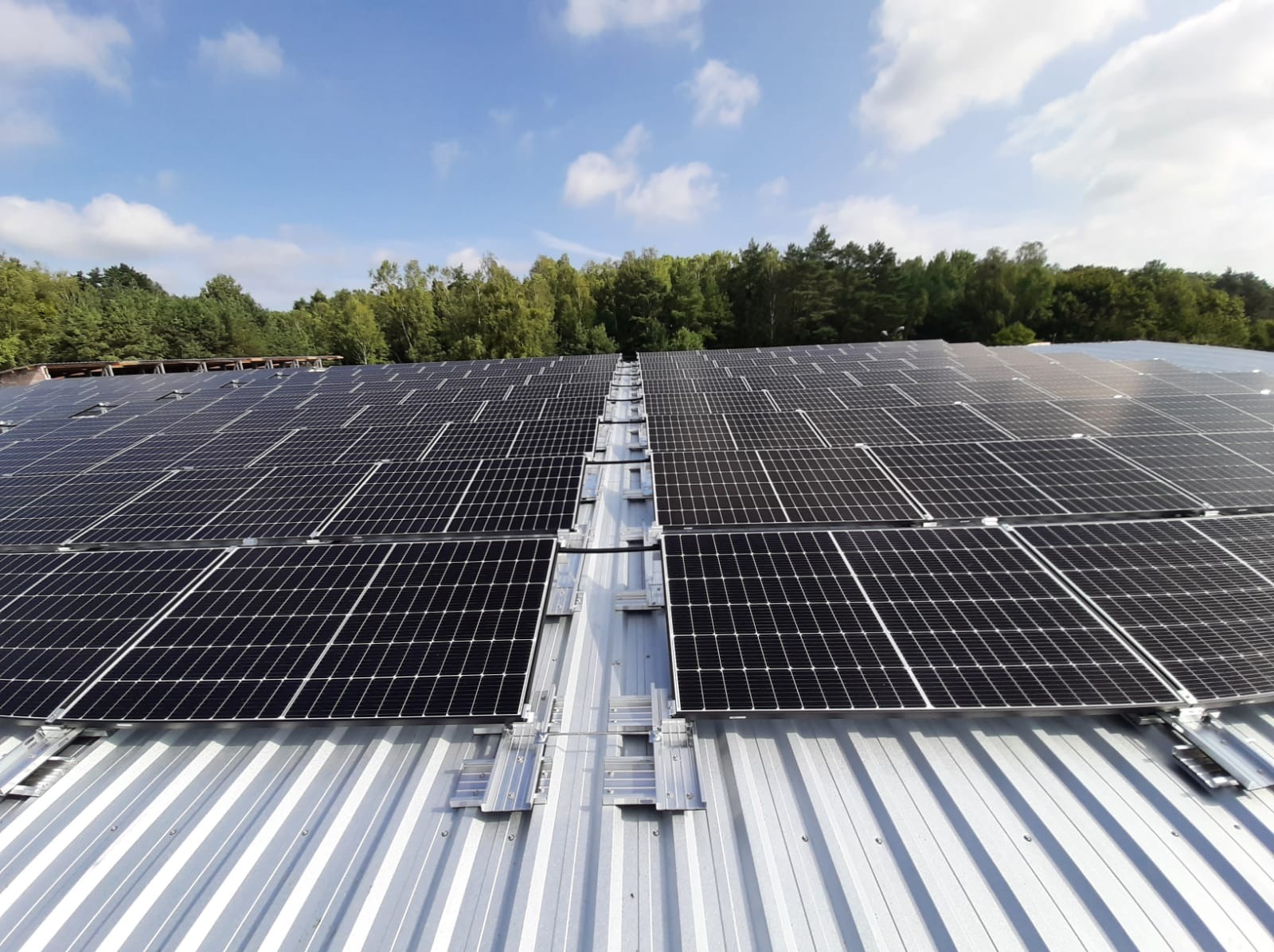 Rozległa instalacja fotowoltaiczna na metalowym dachu budynku przemysłowego, widok z góry, zalesione tło pod błękitnym niebem z chmurami.