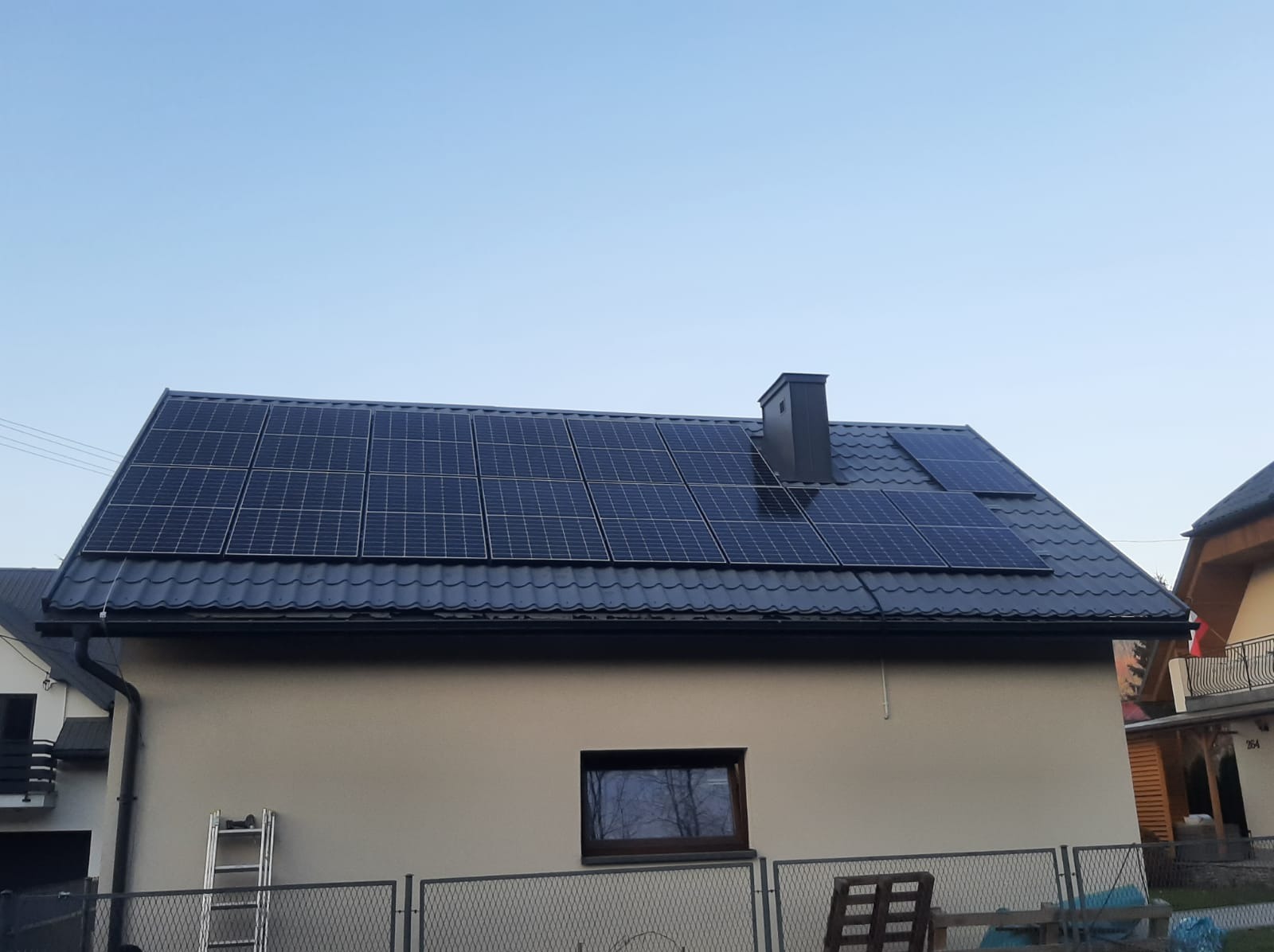 Dom jednorodzinny z panelami słonecznymi na grafitowym dachu z blachy trapezowej, widoczne okno z odbiciem drzew i drabina oparta o ścianę.