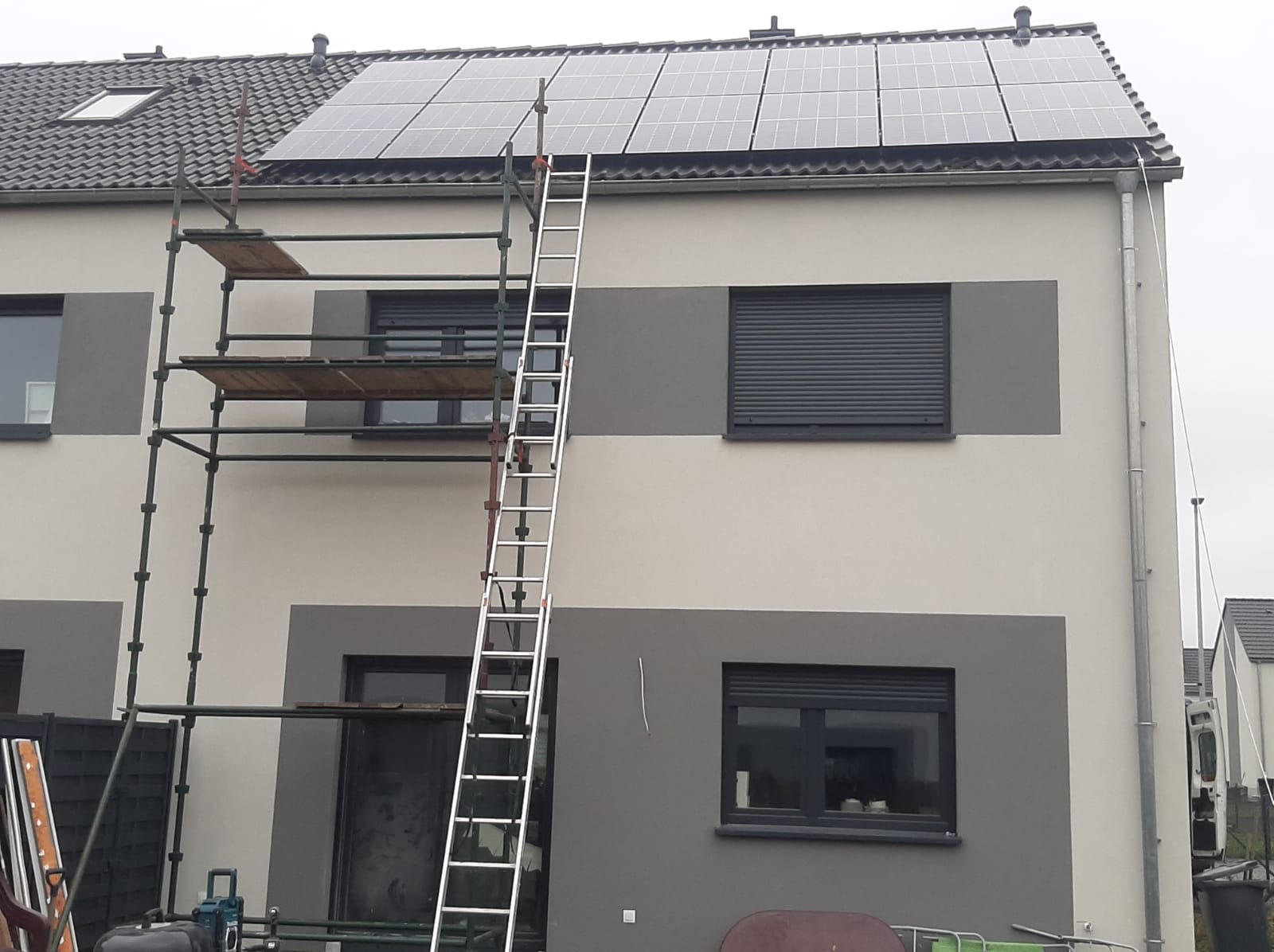 Dwukondygnacyjny dom szeregowy z panelami słonecznymi na dachu, rusztowaniem i drabiną opartą o ścianę, widok z dołu w pochmurny dzień.