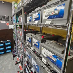 Półki regałowe wypełnione akumulatorami samochodowymi różnych marek i pojemności, z widocznymi cenami i specyfikacjami na etykietach.