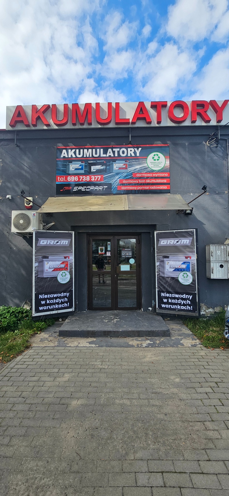 Fasada budynku z szyldem 'AKUMULATORY' i reklamami akumulatorów marki GROM, widoczny numer telefonu i logo SPECPART, wejście do sklepu.