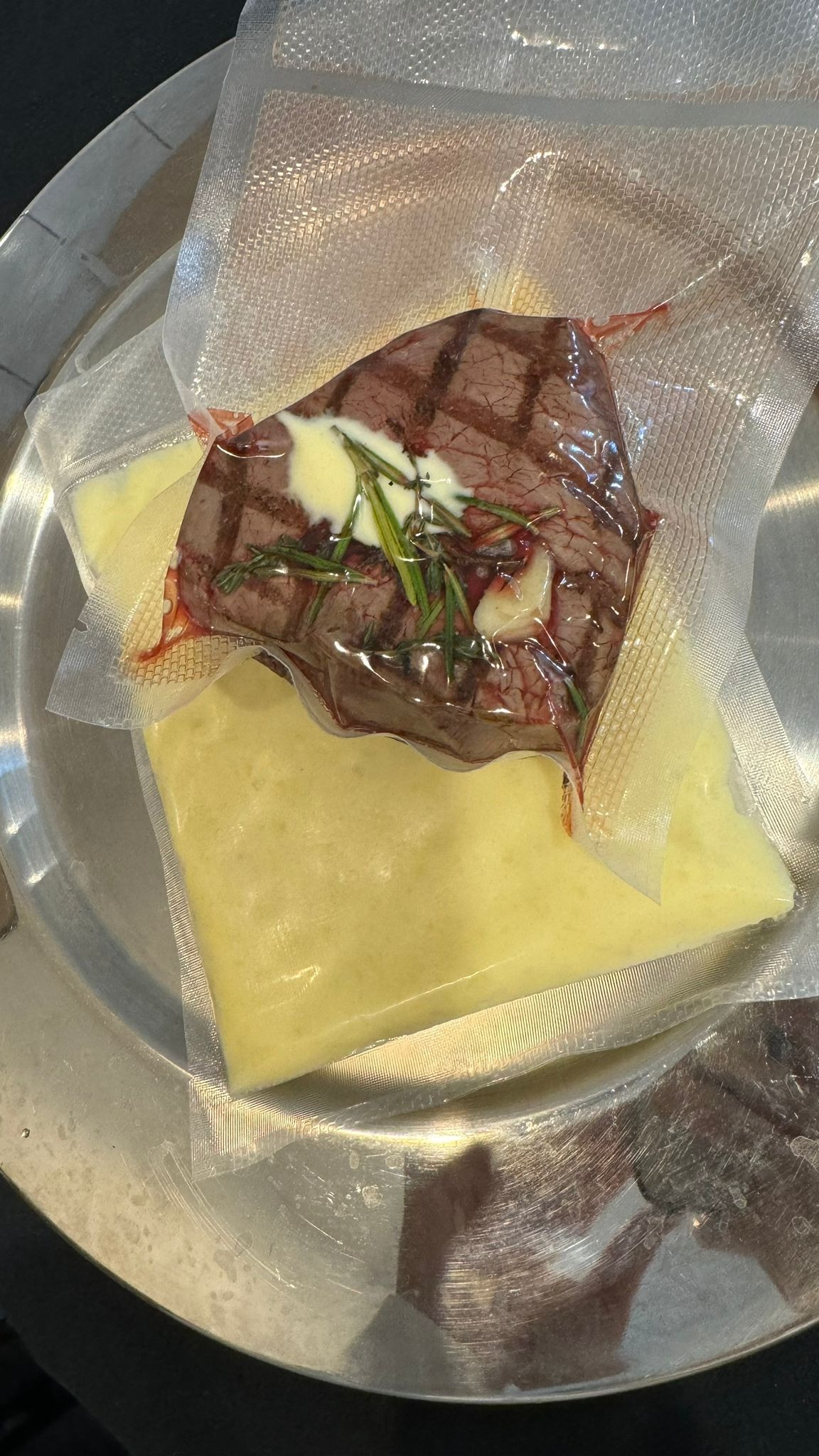 Stek z rozmarynem i sosem, zapakowany próżniowo, leżący na drugim pakunku próżniowym z sosem, na metalowej tacy.