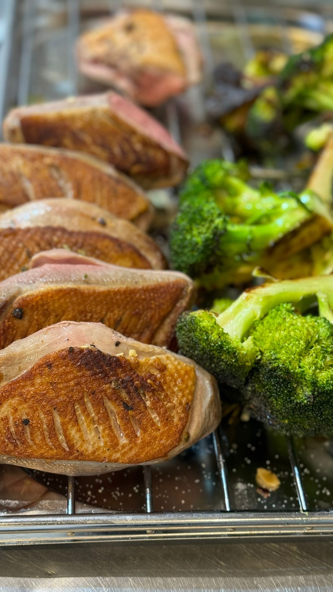 Filety z kaczki z chrupiącą skórką, ułożone obok grillowanych brokułów na metalowej kratce.