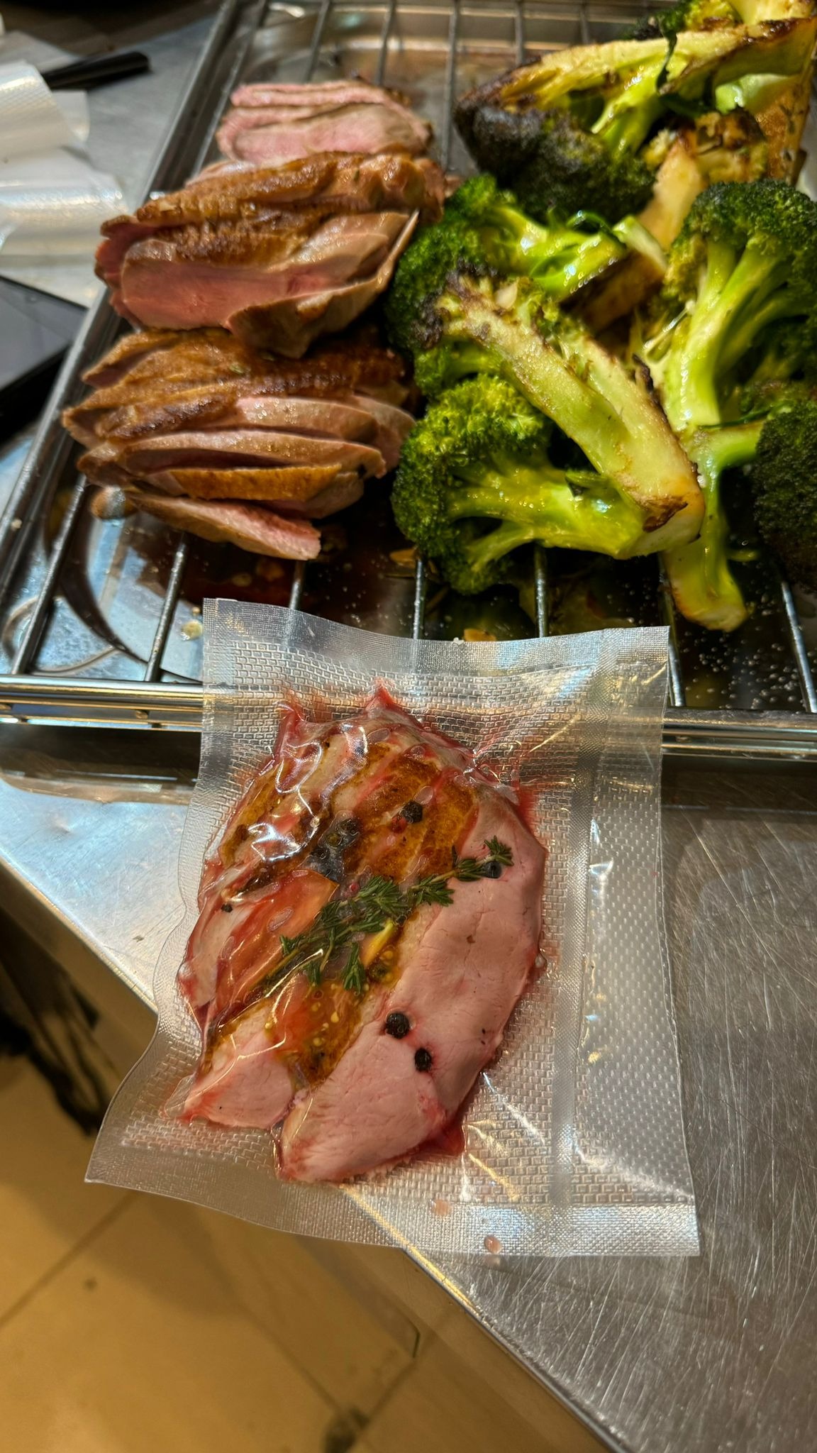 Filet z kaczki sous vide w przezroczystym opakowaniu, obok grillowana brokuła i pokrojone mięso na metalowej kratce.