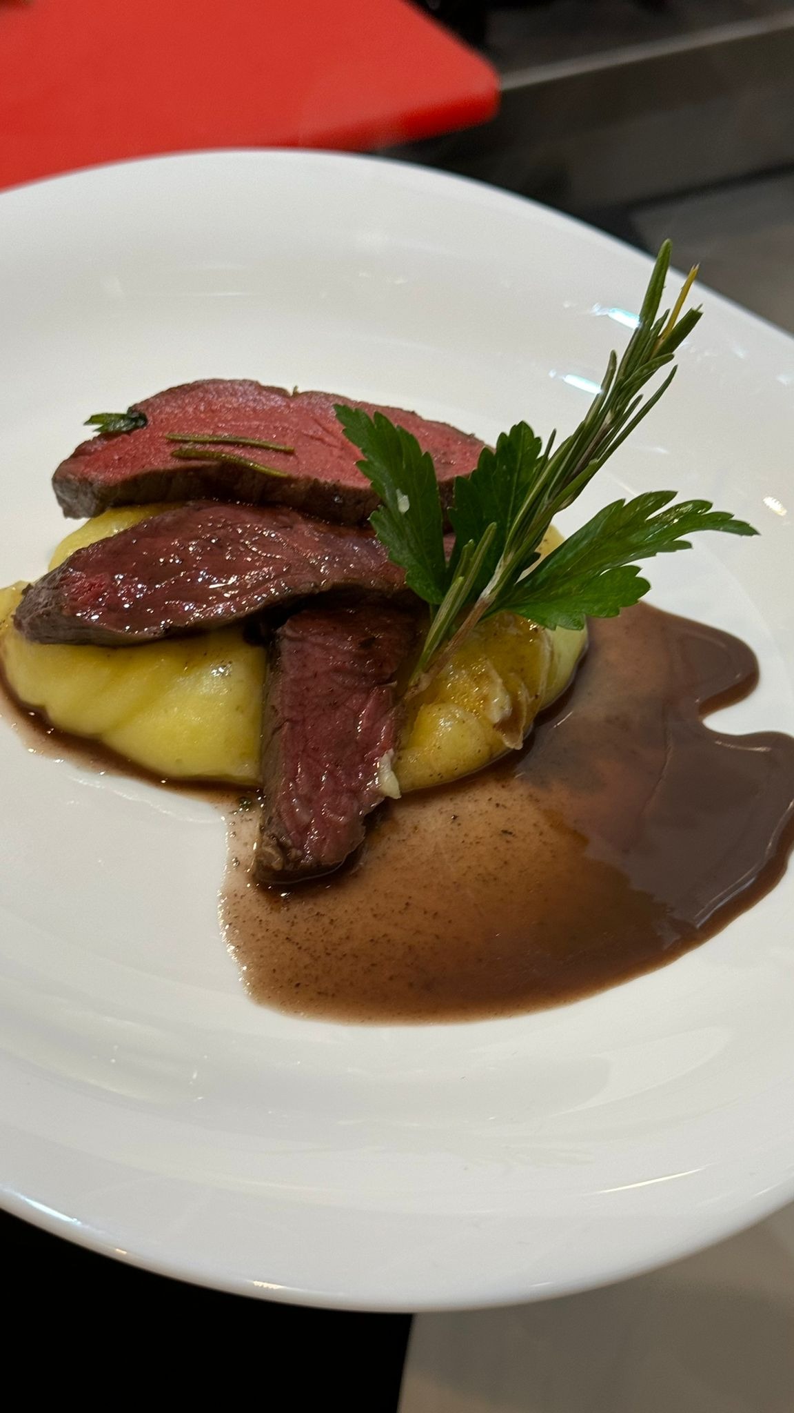 Elegancko podane danie: plastry średnio wysmażonej wołowiny ułożone na purée ziemniaczanym, polane sosem demi-glace i udekorowane gałązką rozmarynu i listkami pietruszki na białym talerzu.