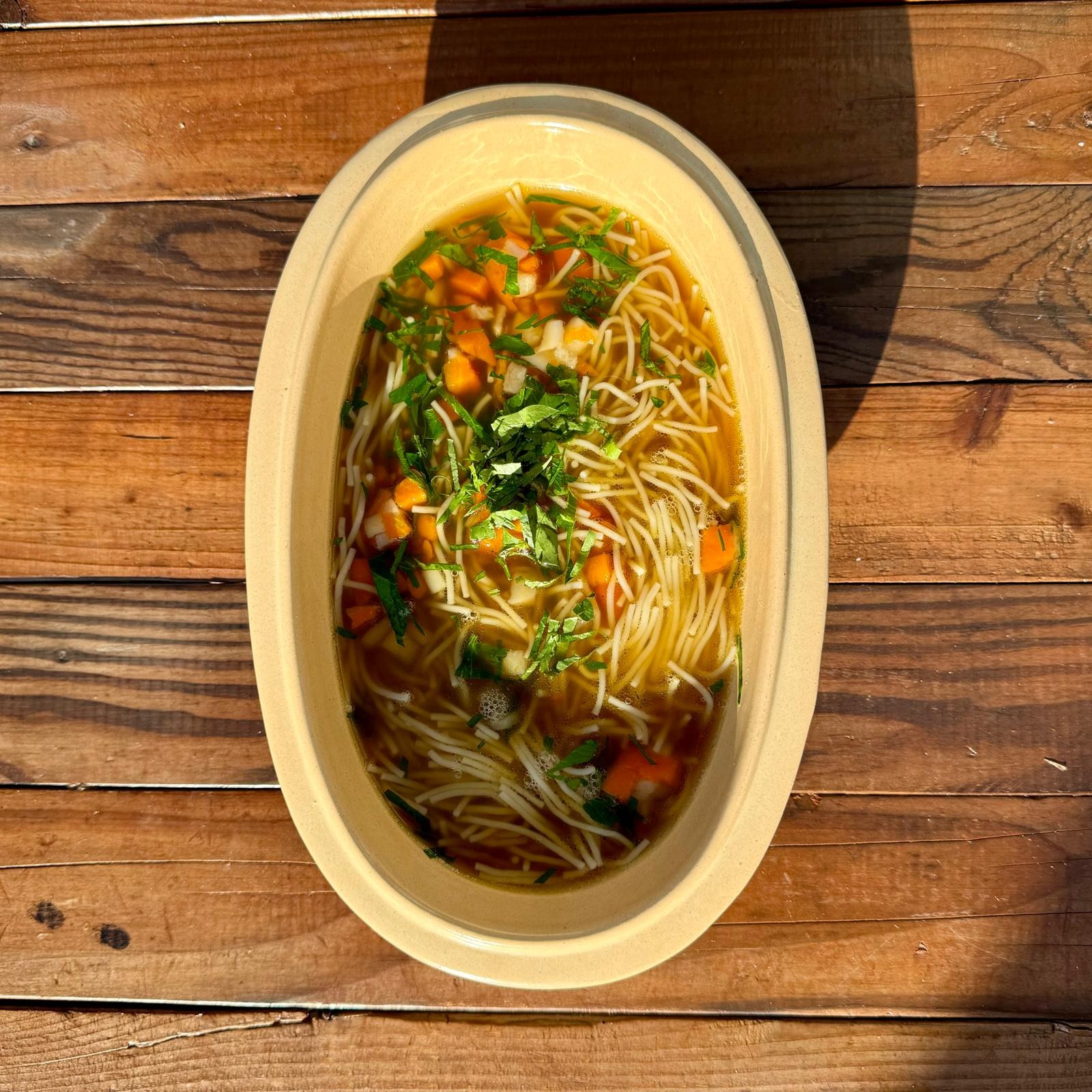 Rosół z makaronem nitki, marchewką, pietruszką i zieloną natką w beżowej, owalnej wazie na drewnianym stole w jasnym, naturalnym świetle.