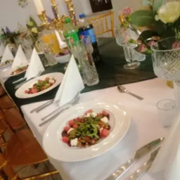 Uroczysty stół z białym obrusem, nakryty talerzami z sałatką z arbuza, sera feta i rukoli, kryształowymi szklankami, złotymi świecznikami i bukietami kwiatów, napoje w butelkach.