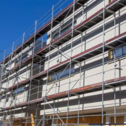 Budynek w trakcie ocieplania elewacji, widoczne rusztowania i zabezpieczone okna folią, ściana pokryta jasnym tynkiem.