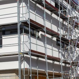 Blue Synergy - Biała elewacja budynku w trakcie prac wykończeniowych, widoczne rusztowania z drewnianymi podestami, okna z szarymi ramami.