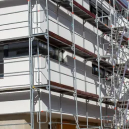 Biała elewacja budynku w trakcie prac wykończeniowych, widoczne rusztowania z drewnianymi podestami, okna z szarymi ramami.