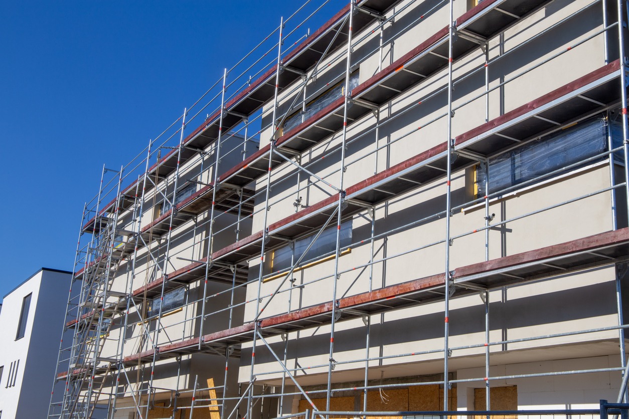 Budynek w trakcie ocieplania elewacji, widoczne rusztowania i zabezpieczone okna folią, ściana pokryta jasnym tynkiem.