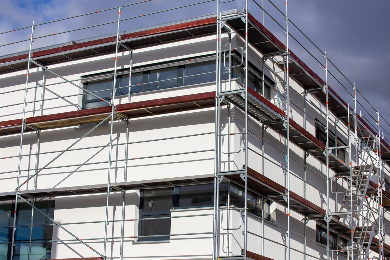 Świeżo otynkowana, biała elewacja budynku w trakcie prac wykończeniowych, z widocznym rusztowaniem i oknami z ciemnymi ramami.