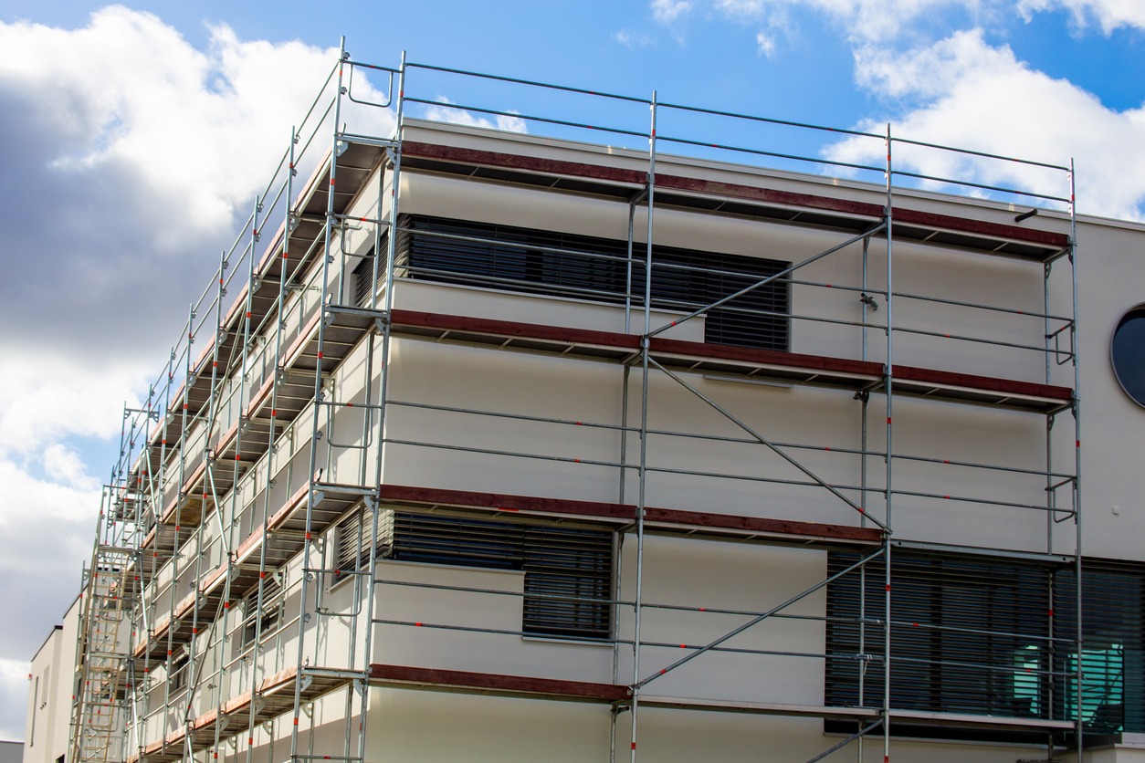 Budynek w trakcie prac elewacyjnych, widoczne rusztowania, biała elewacja i okrągłe okno, błękitne niebo z chmurami w tle.