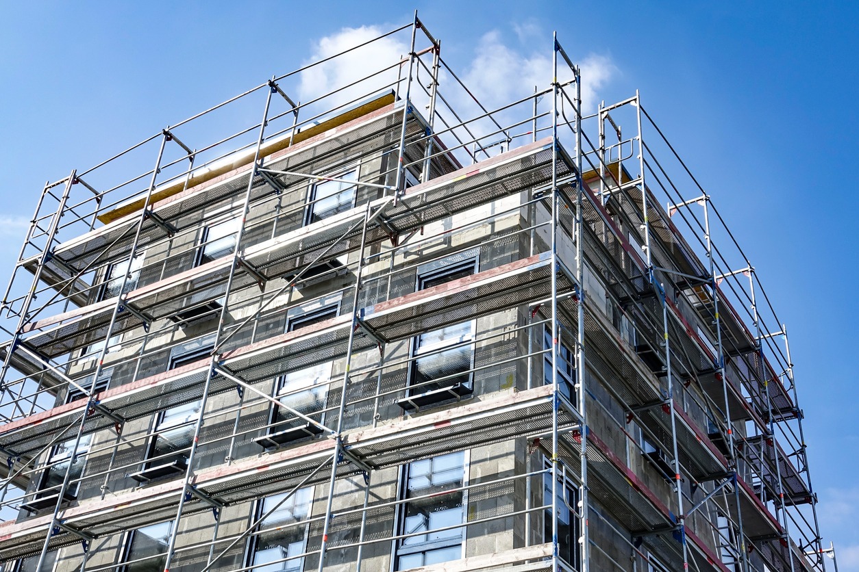 Wielopiętrowy budynek w trakcie prac elewacyjnych, otoczony rusztowaniami na tle błękitnego nieba z delikatnymi chmurami.