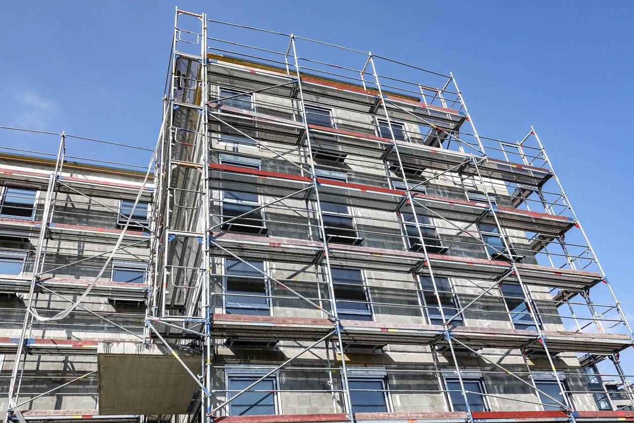 Wielopiętrowy budynek w trakcie prac elewacyjnych z widocznymi rusztowaniami i surową, szarą elewacją przygotowaną pod dalsze prace wykończeniowe.