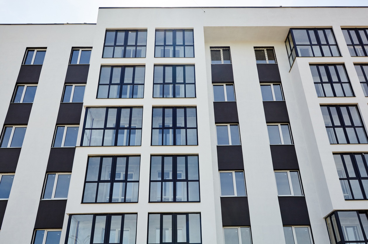 Fragment elewacji nowoczesnego apartamentowca z białymi ścianami i ciemnymi wstawkami, widoczne liczne okna z czarnymi ramami, ujęcie z dołu.