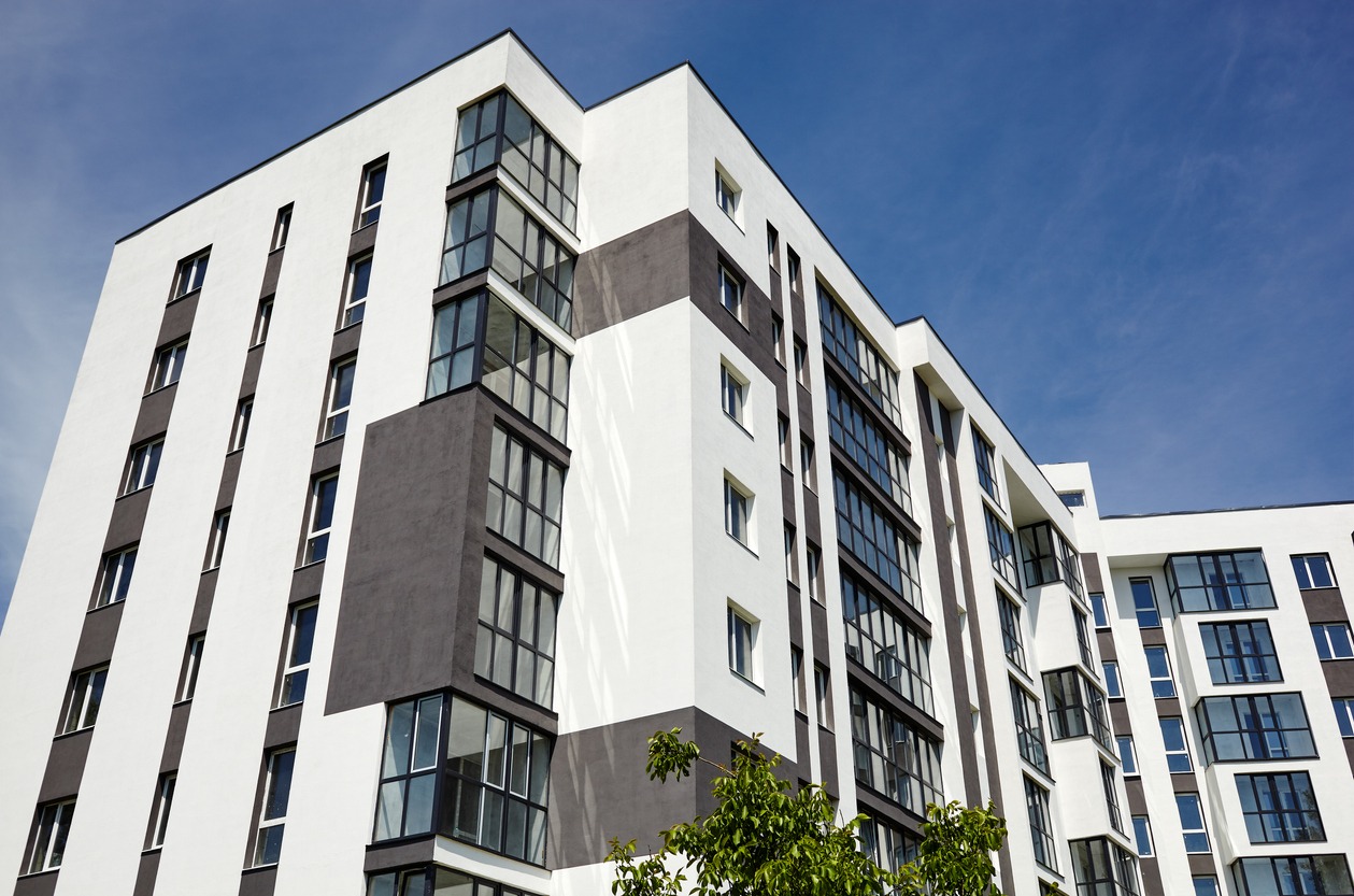 Nowoczesny budynek wielorodzinny z elewacją w kolorach bieli i szarości, z licznymi oknami i balkonami, widziany z dołu na tle błękitnego nieba.