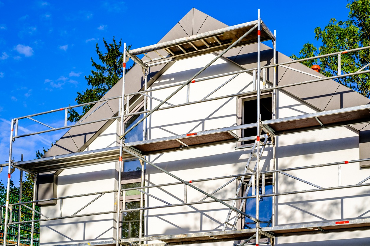 Dom jednorodzinny z białymi ścianami i szarym dachem, otoczony rusztowaniem, na tle błękitnego nieba z fragmentami zieleni.