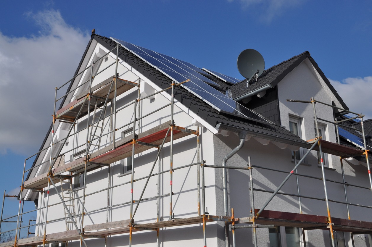 Dom jednorodzinny z białymi ścianami i rusztowaniem, panele słoneczne na dachu, antena satelitarna, błękitne niebo z chmurami w tle.