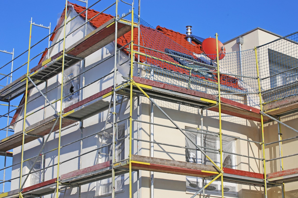 Dom w trakcie prac elewacyjnych, rusztowanie z siatką zabezpieczającą, częściowo zakryte okna folią, widoczny fragment dachu z czerwoną dachówką i instalacją solarną.