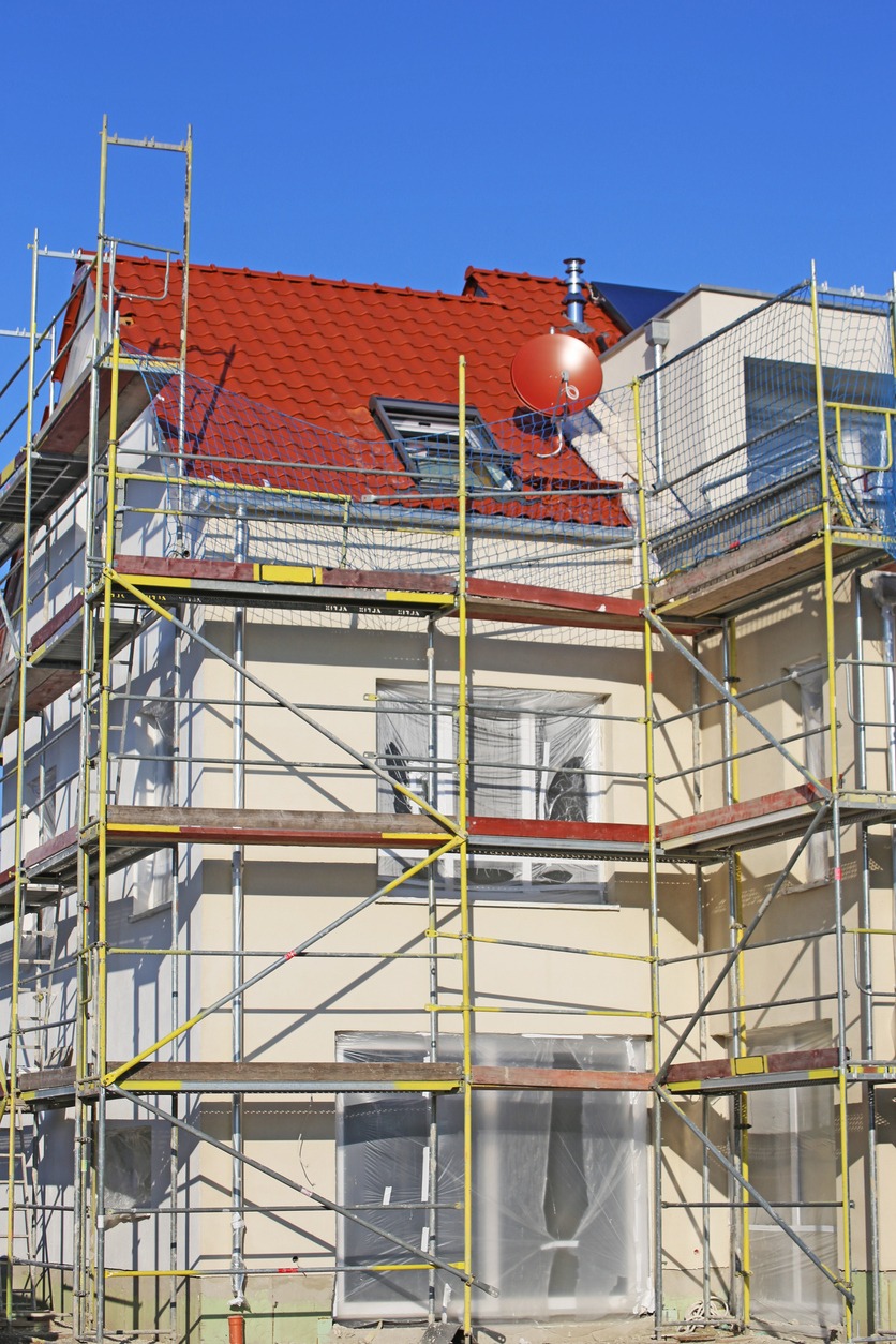 Dom w trakcie prac elewacyjnych, osłonięte okna folią, rusztowanie w kolorze żółtym, czerwony dach z anteną satelitarną.