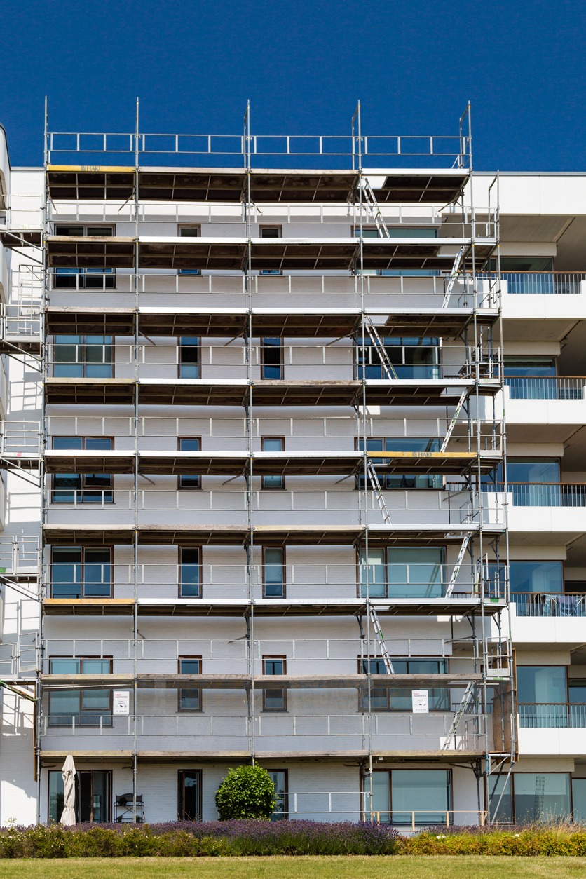 Wielopiętrowy budynek mieszkalny w trakcie prac elewacyjnych, osłonięty rusztowaniem, widoczne okna i balkony, zieleń u podnóża budynku.