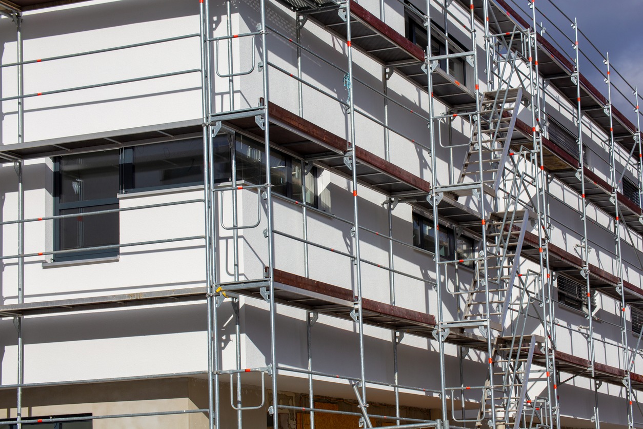 Biała elewacja budynku w trakcie prac wykończeniowych, widoczne rusztowania z drewnianymi podestami, okna z szarymi ramami.