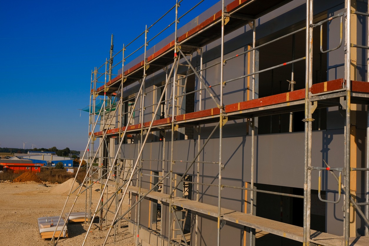 Budynek w trakcie prac elewacyjnych, widoczne rusztowania i częściowo wykończona szara elewacja, w tle budynki przemysłowe i błękitne niebo.