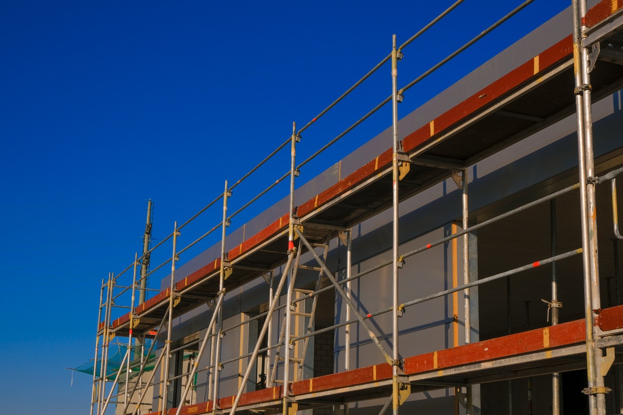 Elewacja budynku w trakcie prac z użyciem rusztowania, widoczne niebieskie niebo i fragment siatki ochronnej.