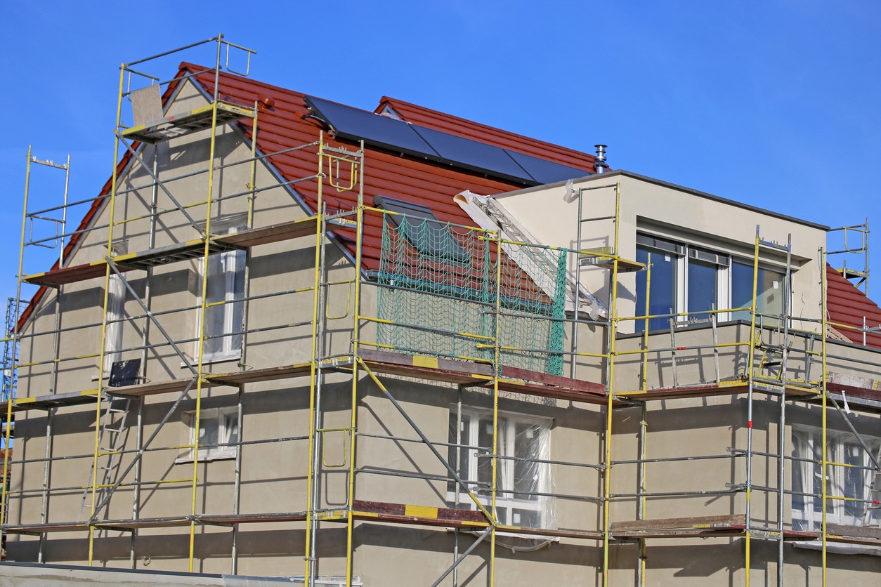 Dom jednorodzinny w trakcie prac elewacyjnych, otoczony rusztowaniem, z widocznymi panelami słonecznymi na dachu i oknami zabezpieczonymi folią.