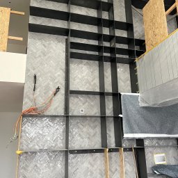 Allconstruct - Ściana wykończona szarą płytką ułożoną we wzór jodełki, z zamontowaną czarną, metalową konstrukcją regału w trakcie montażu, widoczne wystające przewody elektryczne i zabezpieczenia z folii.