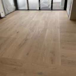 Ułożona drewniana podłoga parkietowa w jasnym odcieniu w nowym domu, duże okna zabezpieczone folią ochronną, widoczne drewniane kliny pod oknami.