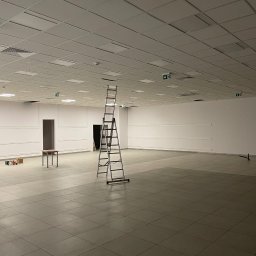 Allconstruct - Przestronne, puste pomieszczenie biurowe z podwieszanym sufitem kasetonowym i białą ścianą, z rozstawioną drabiną aluminiową na środku, stolikiem i paczkami przy ścianie oraz widocznymi znakami...