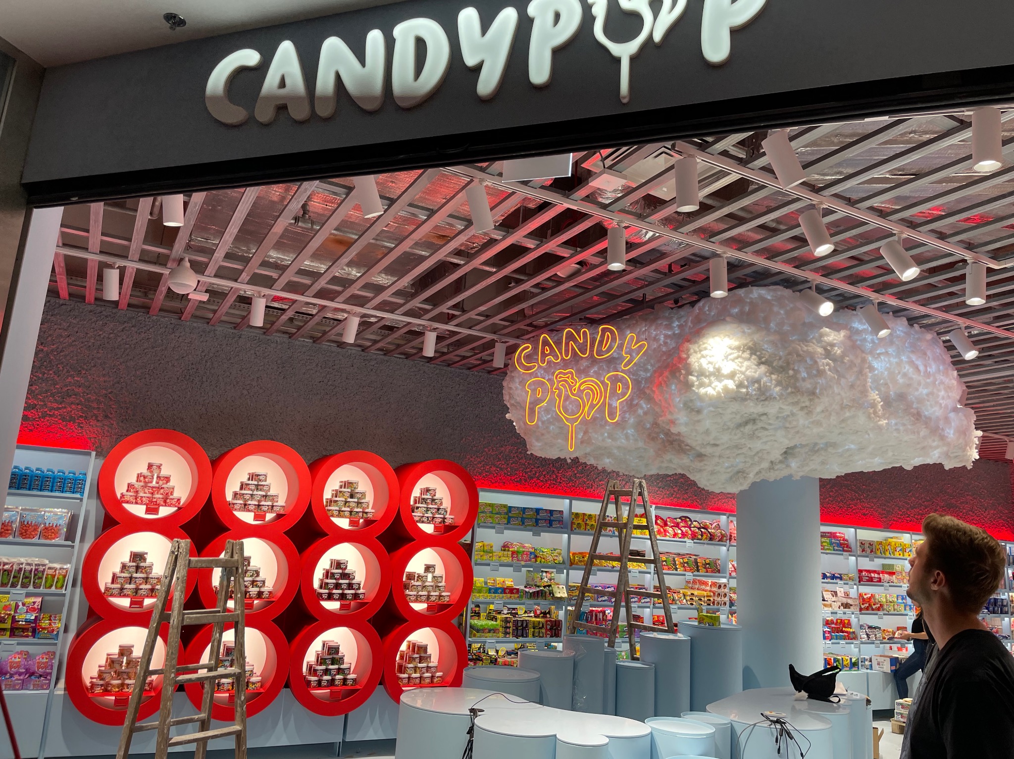 Wnętrze sklepu ze słodyczami z neonowym logo pod sufitem, oświetleniem punktowym i regałami w kształcie czerwonych okręgów, widoczna drabina oparta o regał.