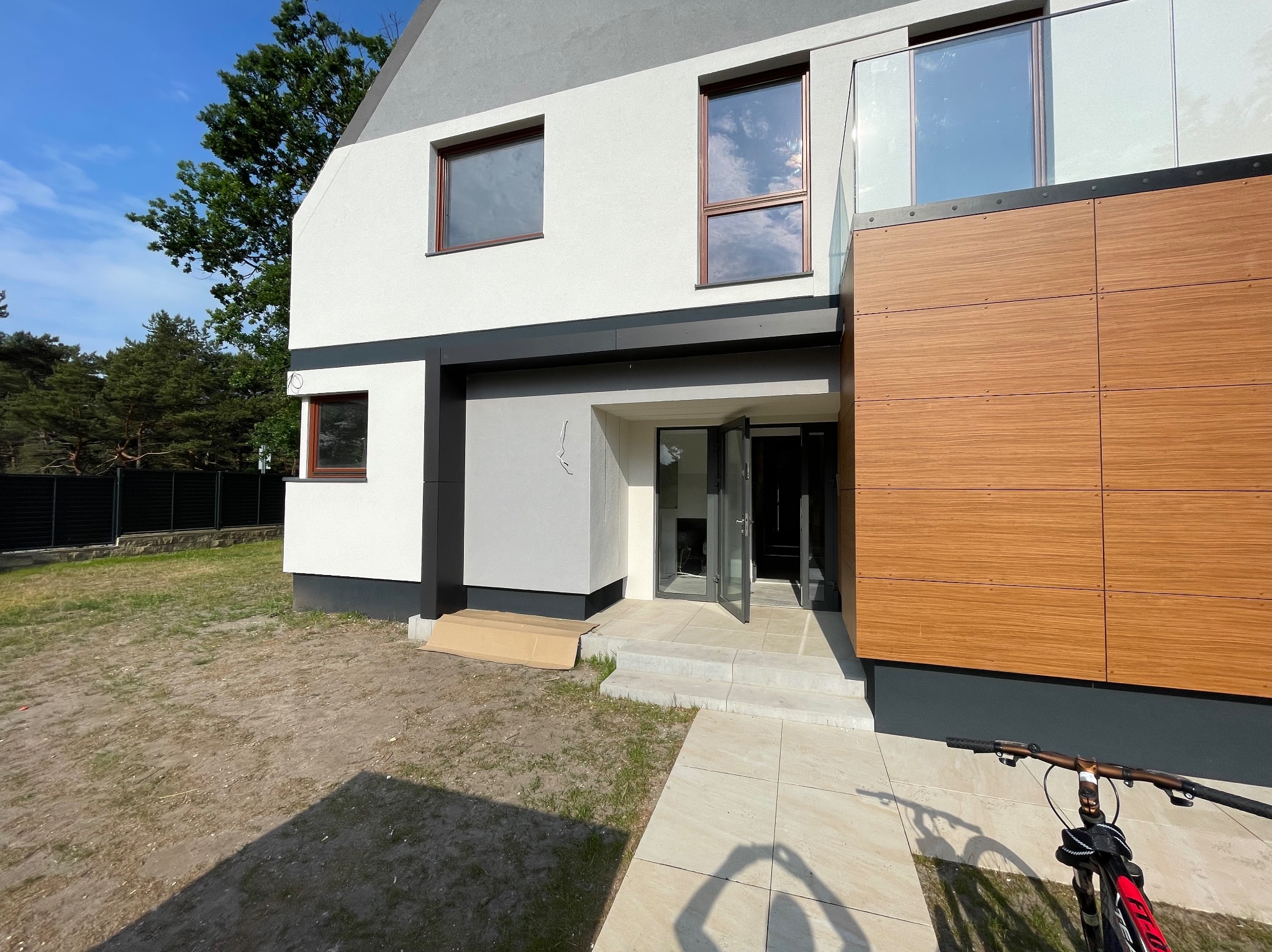 Nowoczesny dom z dwukondygnacyjną elewacją: jasny tynk, drewnopodobne panele, szklana balustrada balkonu, otwarte drzwi wejściowe, karton na schodach, rower w kadrze.