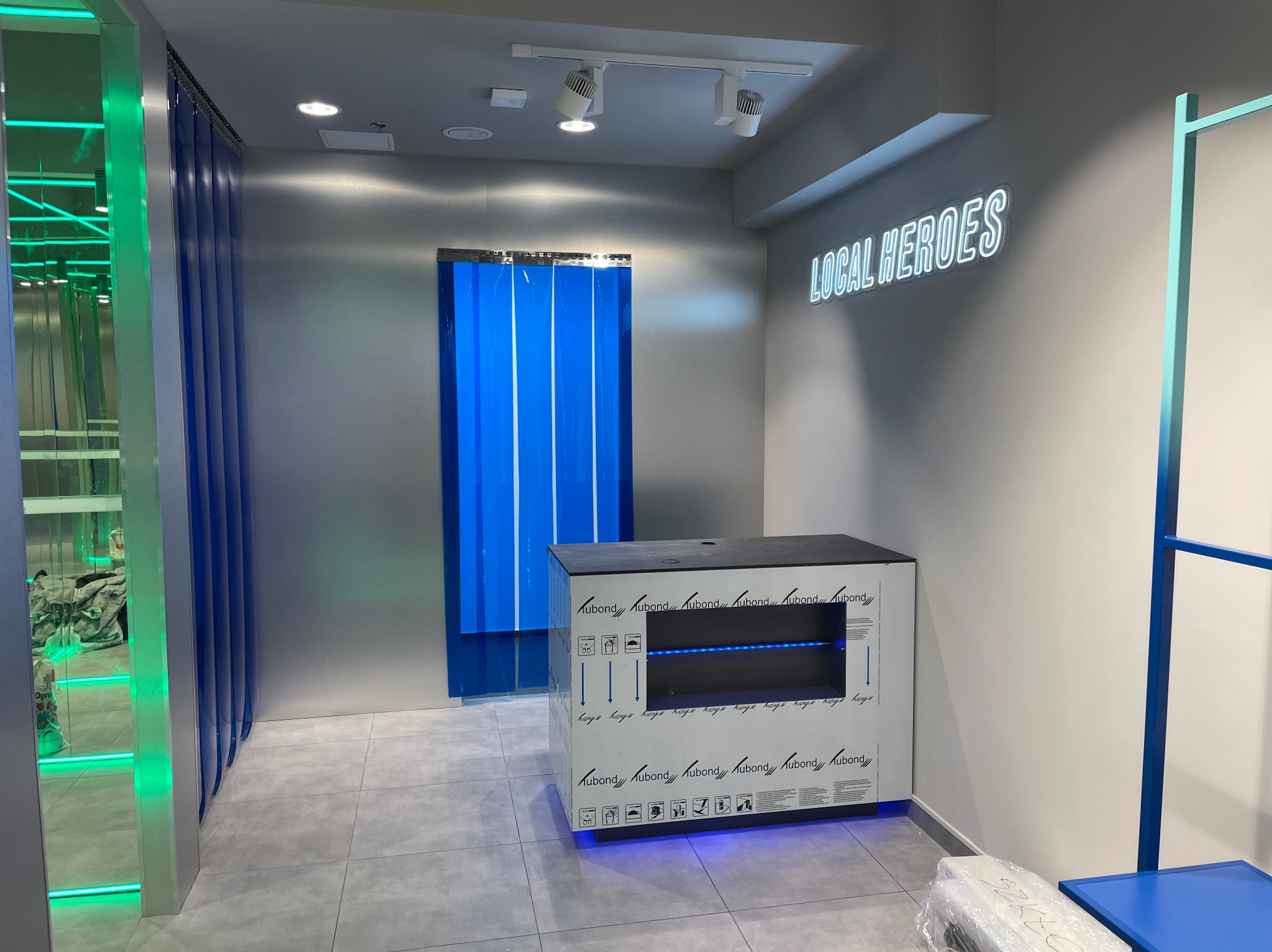 Wnętrze biura po remoncie, ściany w kolorze szarym, neonowy napis 'LOCAL HEROES', kabina z niebieskimi zasłonami, podłoga wyłożona szarymi płytkami, widoczny fragment mebli w kolorze niebieskim.