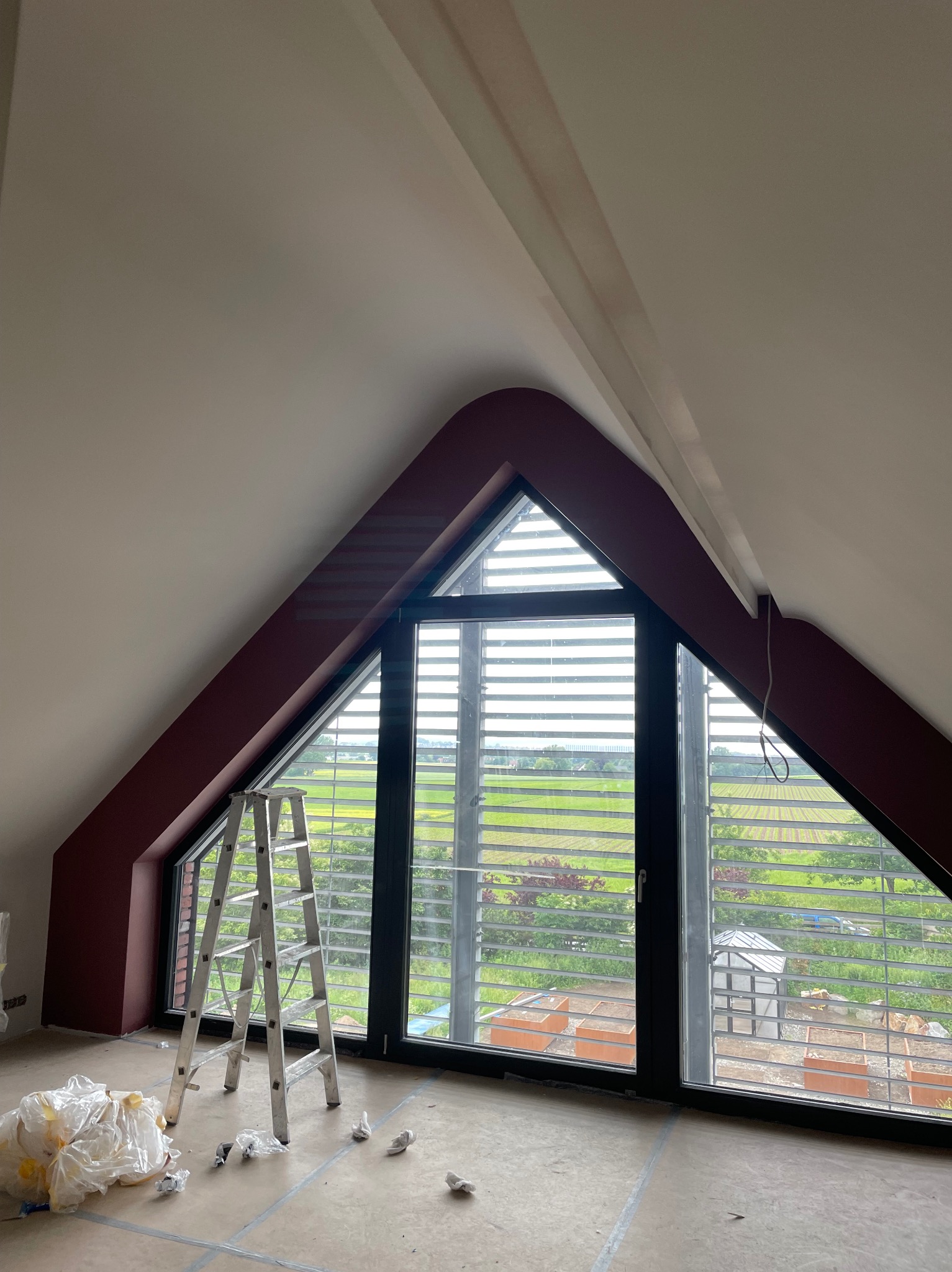 Poddasze z oknem o trójkątnym kształcie w trakcie malowania, widoczna drabina, zabezpieczona podłoga i pomalowane na bordowo obramowanie okna.