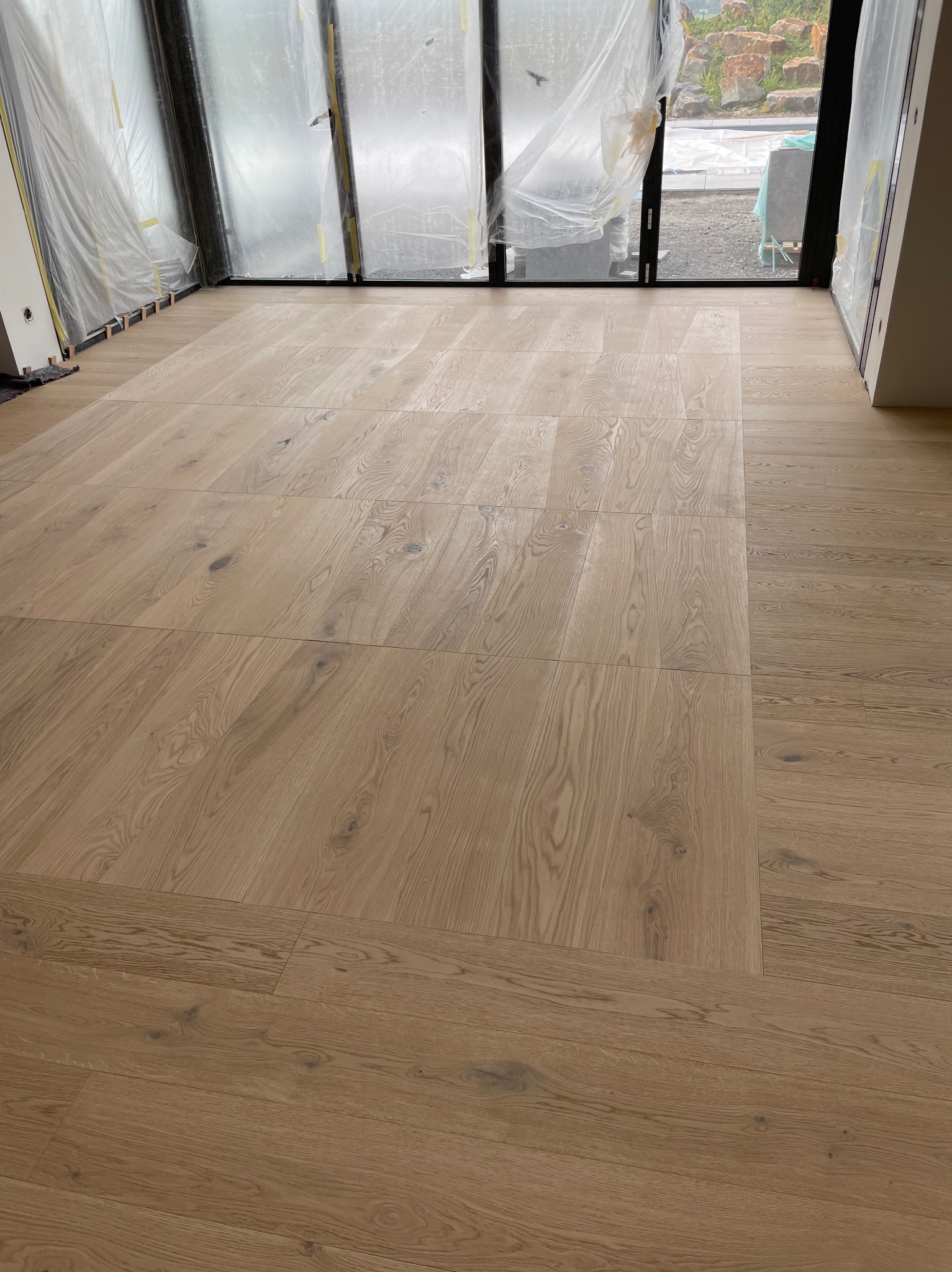 Ułożona drewniana podłoga parkietowa w jasnym odcieniu w nowym domu, duże okna zabezpieczone folią ochronną, widoczne drewniane kliny pod oknami.