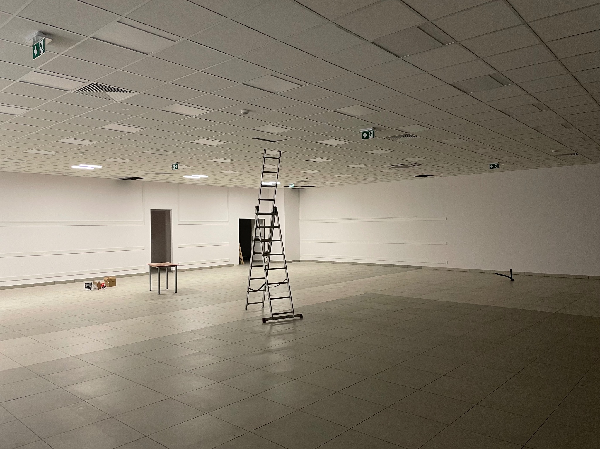 Przestronne, puste pomieszczenie biurowe z podwieszanym sufitem kasetonowym i białą ścianą, z rozstawioną drabiną aluminiową na środku, stolikiem i paczkami przy ścianie oraz widocznymi znakami...