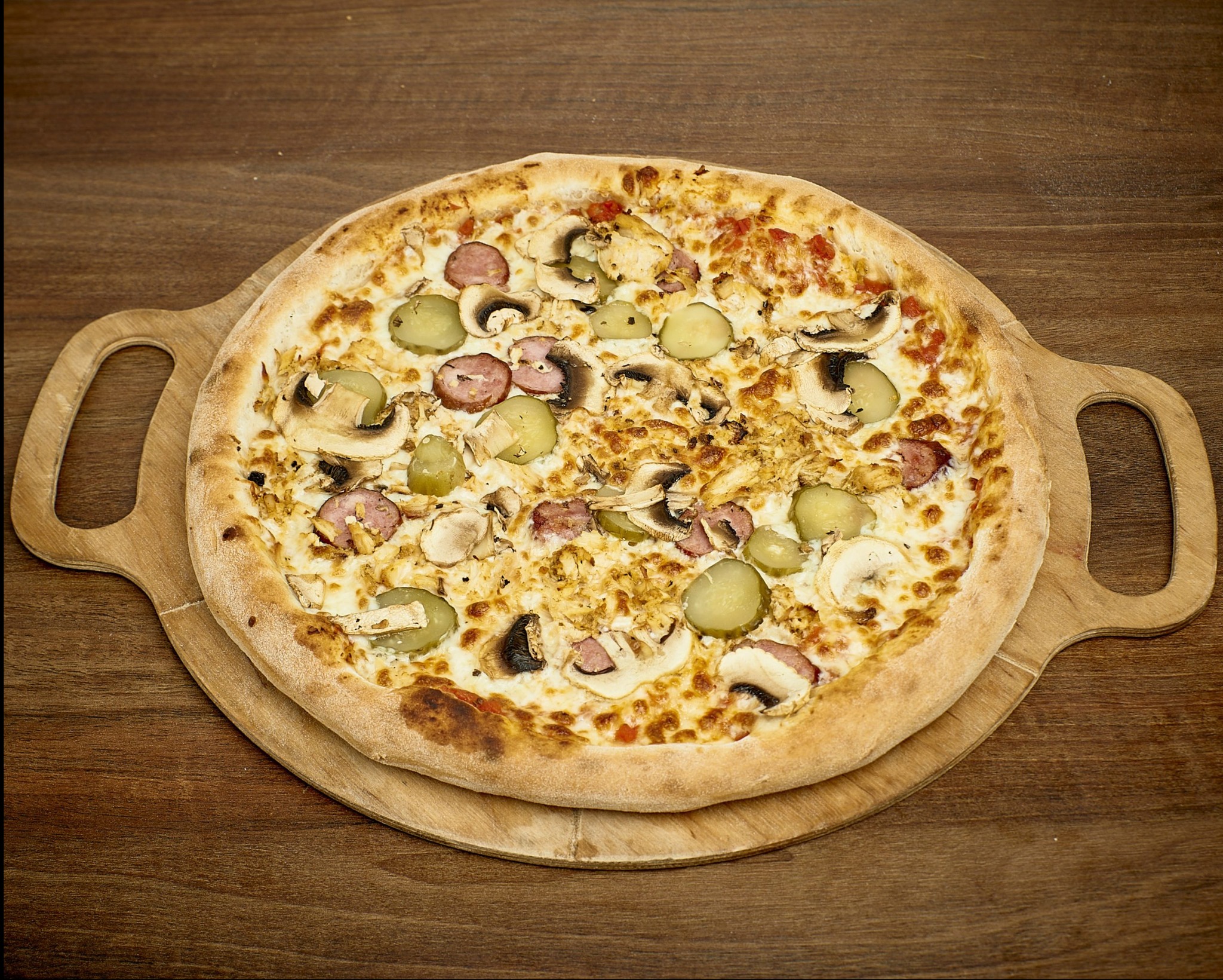 Okrągła pizza z pieczarkami, ogórkiem konserwowym, kawałkami kurczaka i plasterkami kiełbasy na drewnianej desce z uchwytami, na tle drewnianego stołu.