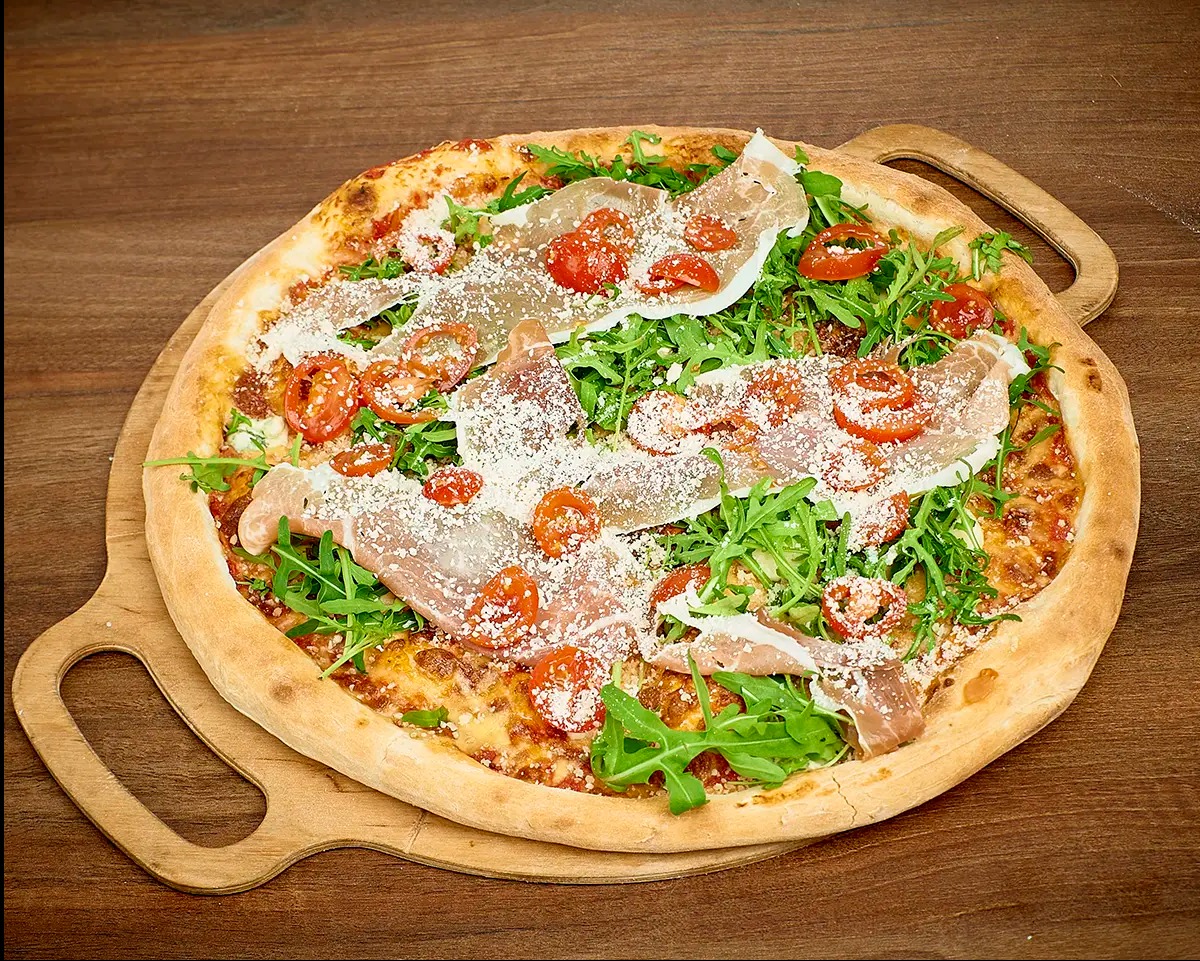 Pizza z szynką parmeńską, rukolą, pomidorkami cherry i parmezanem, podana na drewnianej desce z uchwytami.